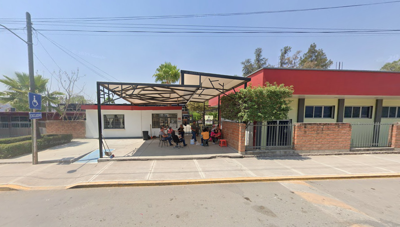 Fotografía en la que se muestra una explanada que está techada. Al finalizar la explanada hay una caseta de color blanco. A la derecha de la caseta está la reja de acceso. El plantel está rodeado por muros de tabique rojo intercalados con rejas de color gris.